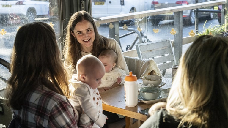 Mums with small babies chatting in the café at Sizergh. They are sitting at a table next to the large floor to ceiling windows, and coffee cups and plates of cake are on the table.